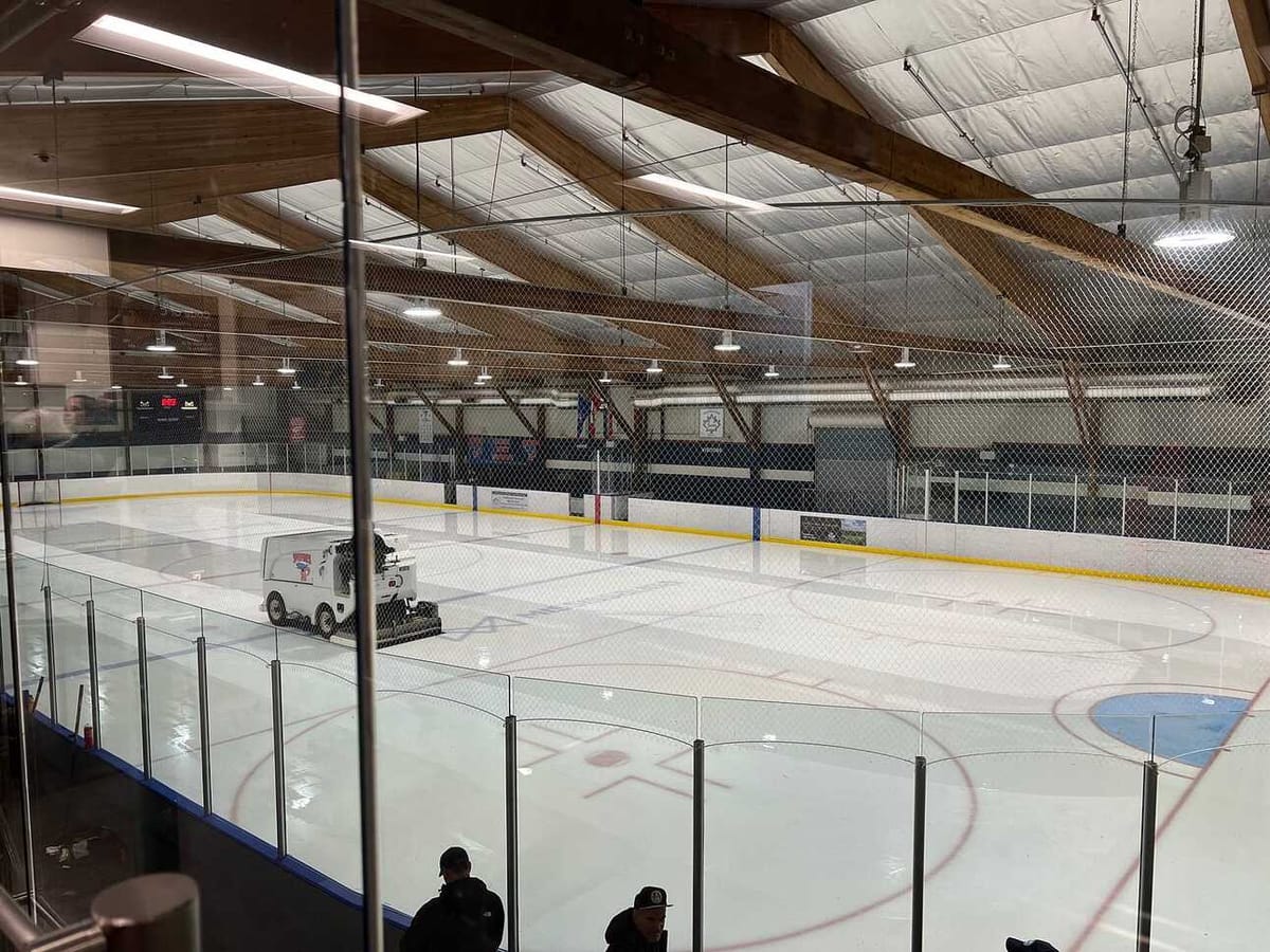 A Zamboni resurfacing the ice in an empty indoor hockey rink, seen through the glass